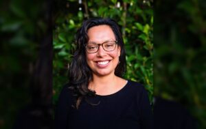 Alicia, wearing black-framed glasses, smiles while looking directly at the camera lens, in front of a wall of greenery.